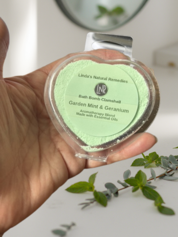 Hand holding a green bath bomb with 'Linda's Natural Remedies' label, surrounded by greenery.