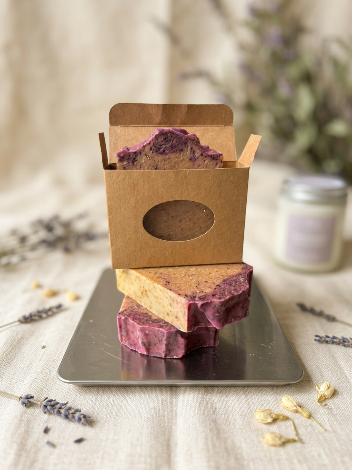 Three soap bars stacked on a metal tray with a brown box on top, surrounded by lavender flowers.
