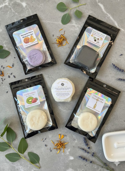 A collection of handcrafted solid shampoo bars from Linda's Natural Remedies, including round and shell-shaped bars in various colors like purple, charcoal, and cream, displayed with natural botanical ingredients.