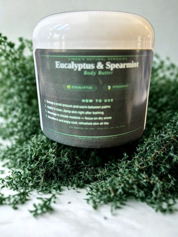 A jar of Linda's Natural Remedies Eucalyptus & Spearmint Body Butter nestled in a bed of fresh, green botanical leaves.