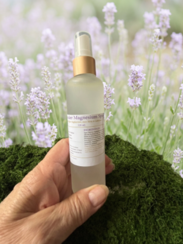 A hand holds a frosted glass spray bottle labeled 'Magnesium Spray' with a gold collar and clear cap, set against a blurred background of lavender flowers and green moss.
