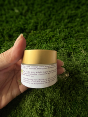 A hand holding a small white cosmetic jar with a gold lid, set against a background of green moss or artificial grass.