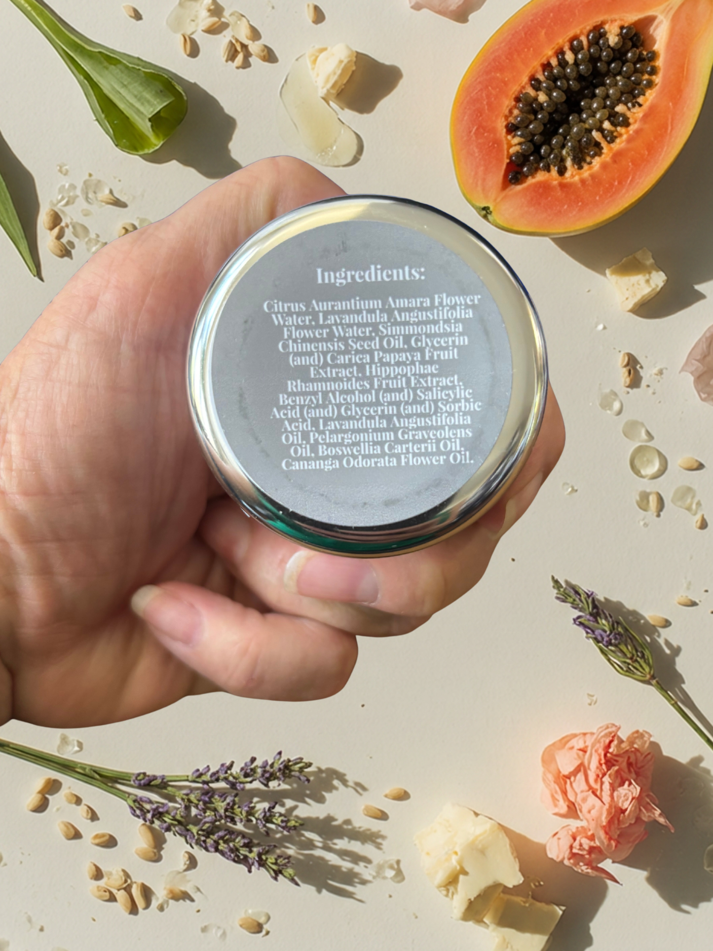 Hand holding a container with ingredients list, surrounded by natural elements like papaya, lavender, and flowers.