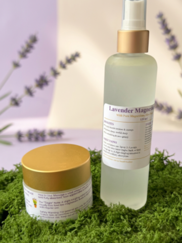 A skincare set featuring a clear spray bottle labeled 'Lavender Magnolia' with a gold pump, and a white jar with a gold lid, both placed on green moss with blurred lavender sprigs in the background.