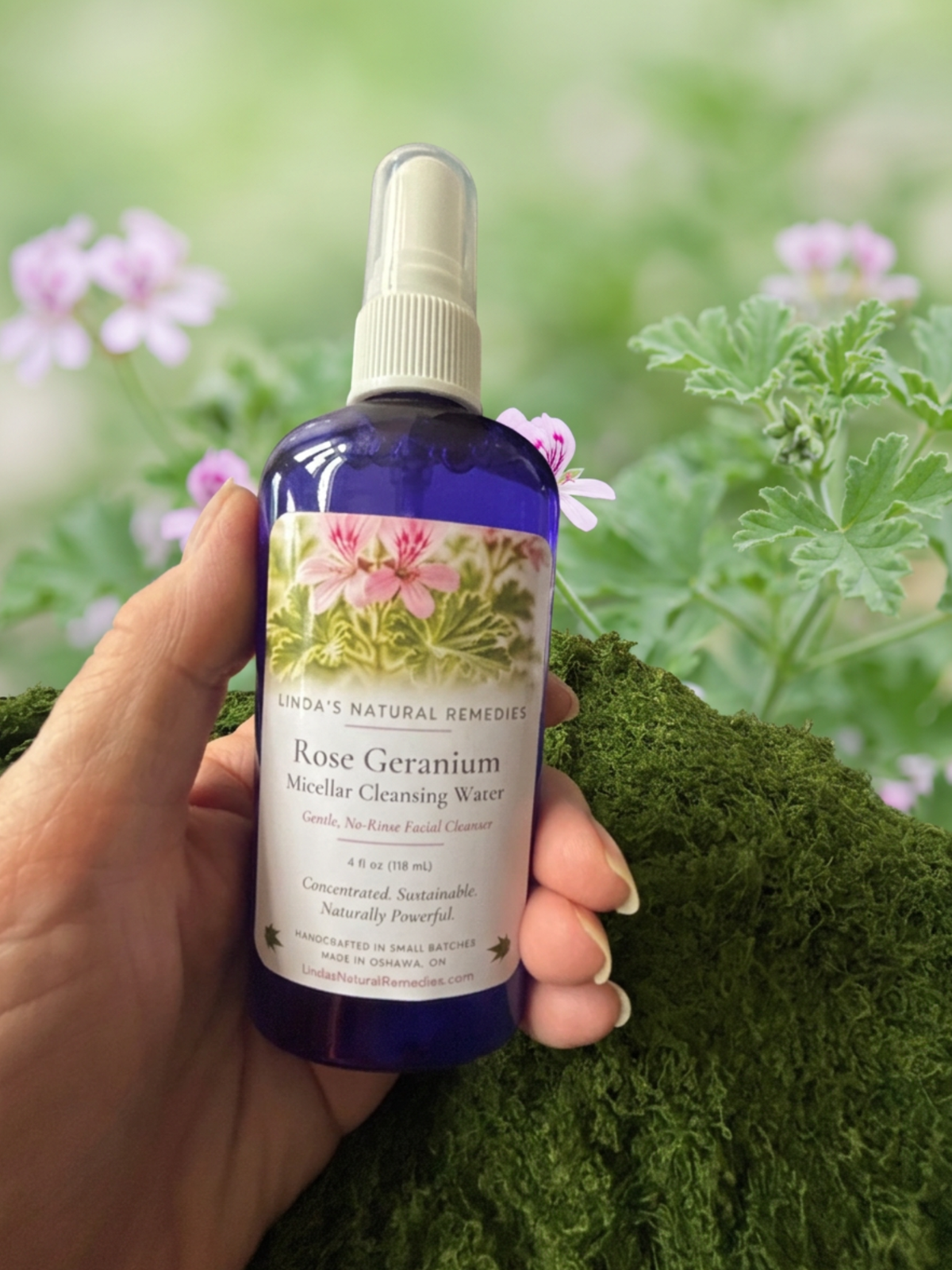 A hand holds a bottle of Linda's Natural Remedies Rose Geranium Micellar Cleansing Water against a blurred green garden background with small pink flowers.