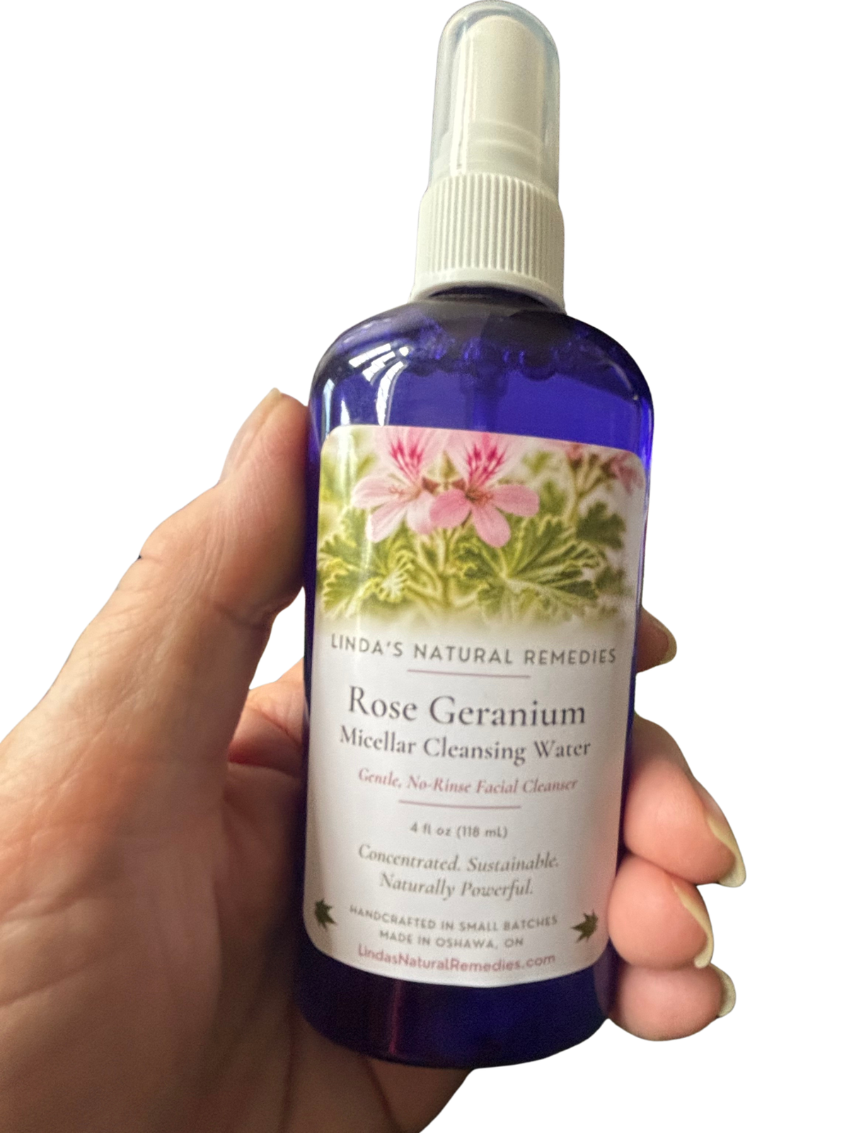 A hand holding a bottle of Linda's Natural Remedies Rose Geranium Micellar Cleansing Water. The bottle is a deep cobalt blue glass with a white spray top and a label featuring pink rose geranium flowers and green leaves.