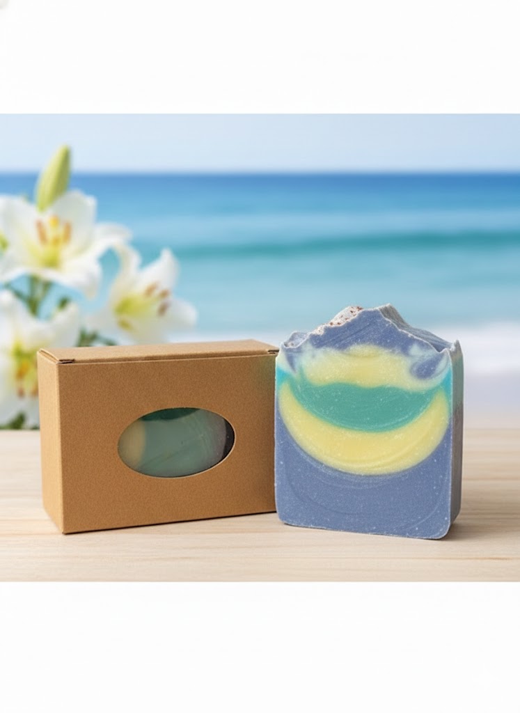 A tall, hand-cut artisan soap bar with a natural, textured top, featuring swirling layers of deep periwinkle, teal, and soft yellow mica. The soap stands next to a brown kraft paper box with an oval window, set against a blurred background of a sunny beach and white lilies.