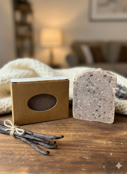 A tall, rustic Vanilla Cashmere artisan soap bar with a speckled cream and earth-tone texture stands next to a kraft paper box with an oval window. The scene is set on a dark wood surface with vanilla beans and a cozy white knit blanket in a warm, blurred living room setting.