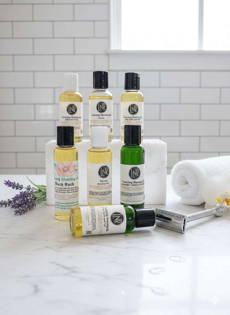 A professional collection shot of six bottles of Linda's Natural Remedies Foaming Shaving Oils arranged on a white marble surface in a bright, modern bathroom. The lineup includes scents like Karma, Sea Salt and Lily, and Lavender, displayed with a chrome safety razor and a soft white towel in the background.