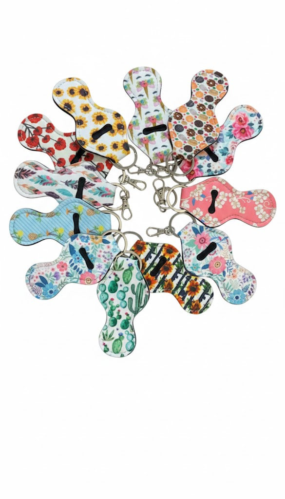 "A variety of fabric lip balm holder keychains in floral and botanical patterns, including sunflowers, cacti, and colorful petals, arranged in a circle on a white background."