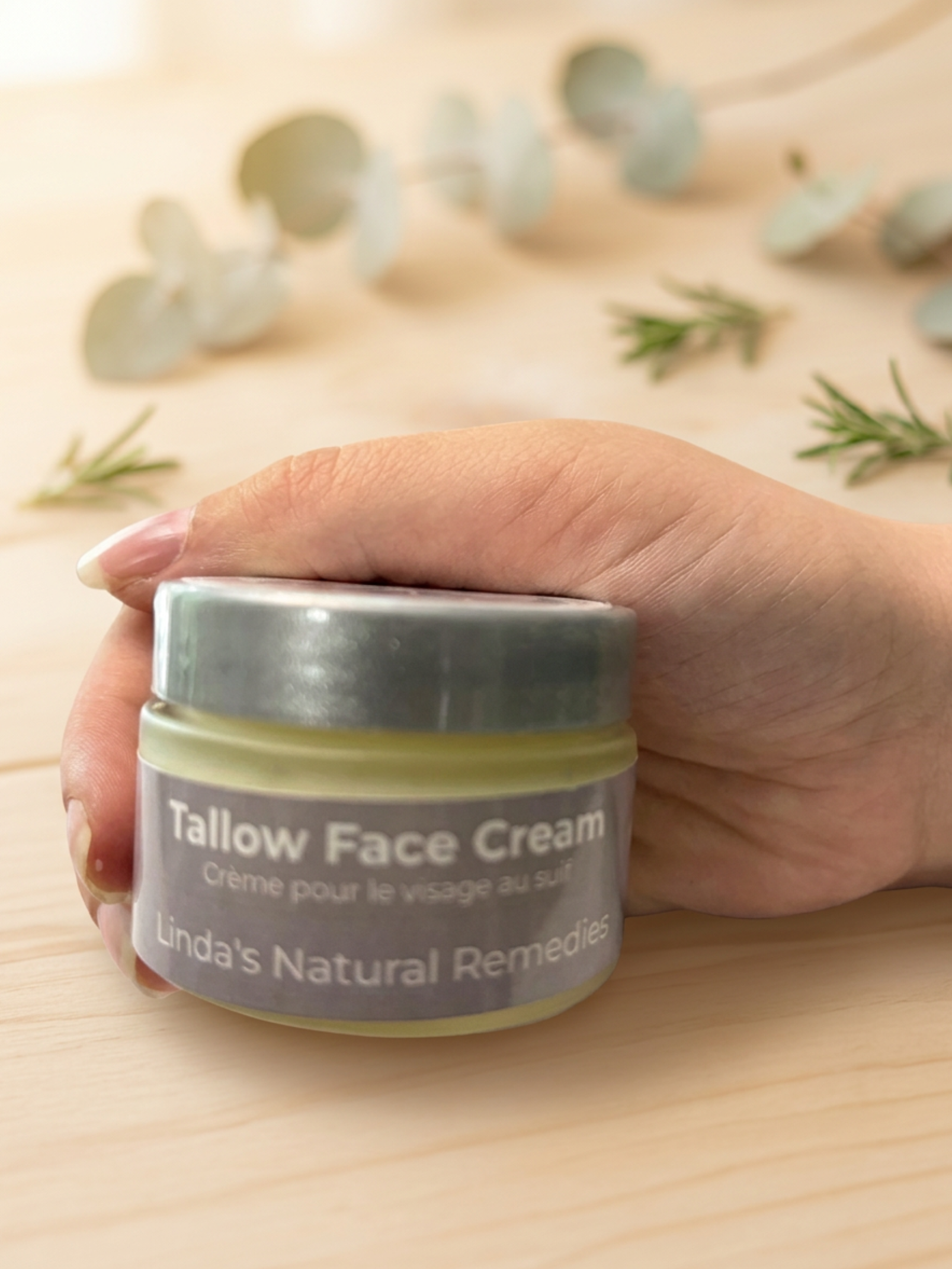 Hand holding a jar of Tallow Face Cream by Linda's Natural Remedies on a wooden surface with eucalyptus leaves.
