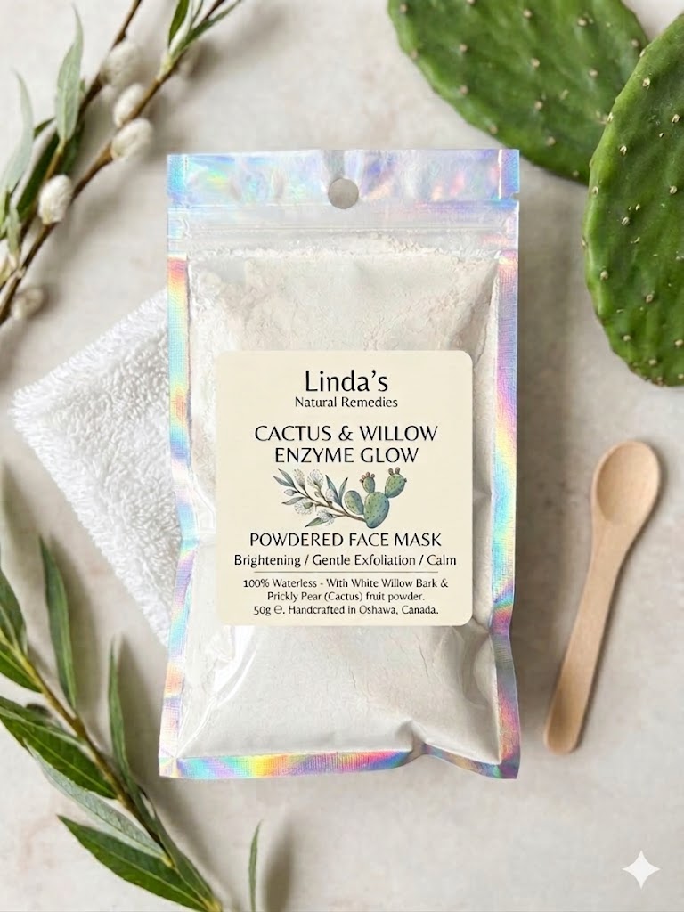 A top-down view of Cactus & Willow Enzyme Glow Powdered Face Mask by Linda’s Natural Remedies in a holographic pouch, paired with a soft white microfiber face cloth, a wooden application spoon, and fresh cactus leaves.